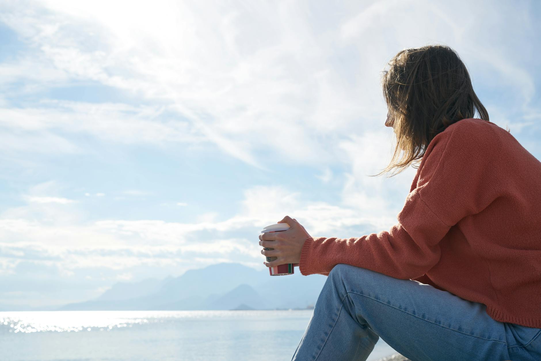 A woman in a cozy orange sweater sits by the water's edge, holding a cup, gazing thoughtfully at the serene landscape featuring mountains and a reflective sea under a cloudy sky.